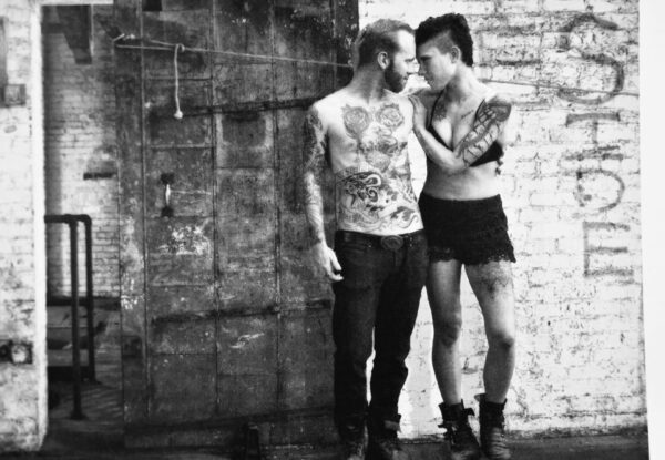 Tattooed couple stand close by brick wall.